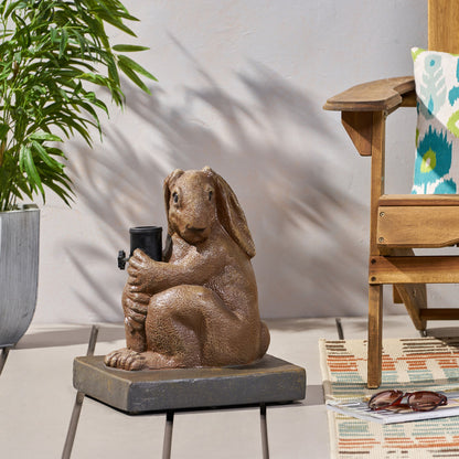 A light brown Hand Crafted Rabbit Umbrella Base with a playful design is placed on a patio deck beside a potted plant, a wooden chair featuring a colorful cushion, and a patterned rug.