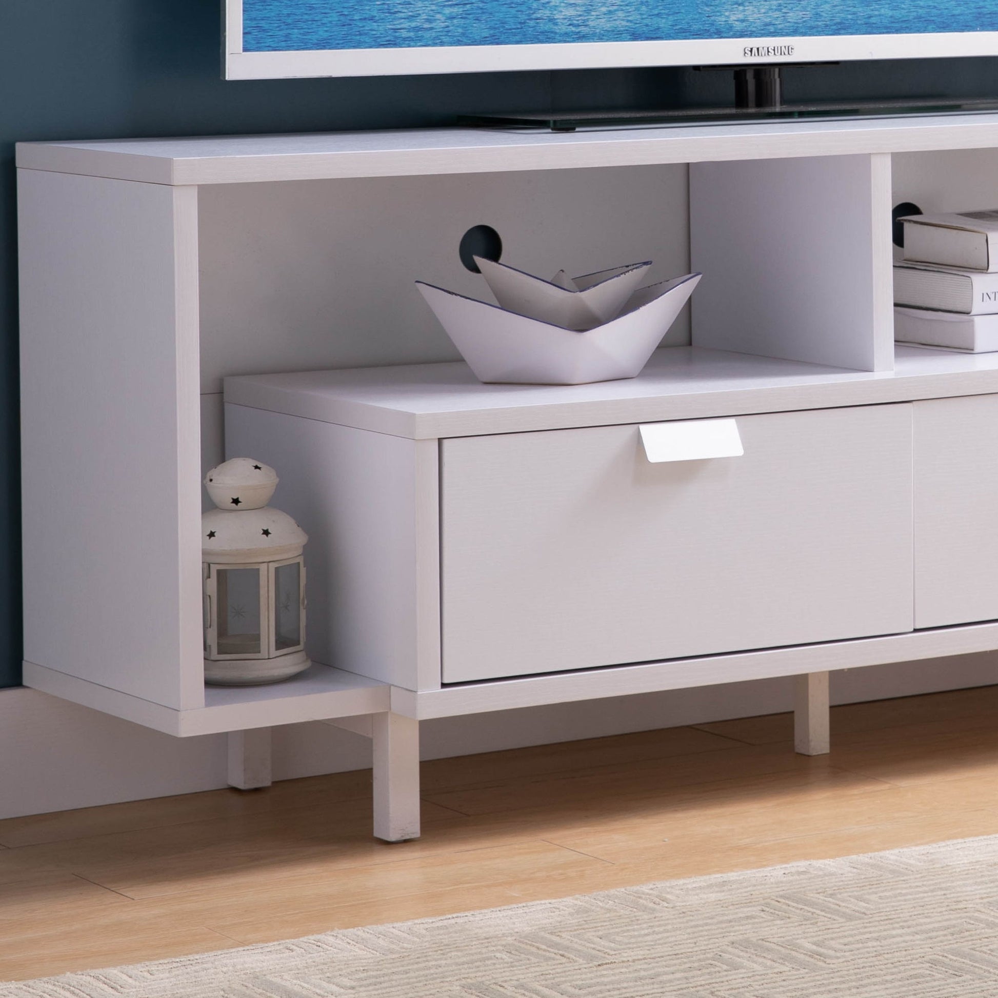 The Modern TV Stand & Two Shelves And Two Drawers stands on a wooden floor near a beige rug. Its shelves hold a lantern, decorative bowl, and books, with a TV partially visible on top.