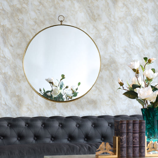 A Round Mirror, Circle Mirror With Iron Frame, reflects white flowers and hangs above a tufted dark gray sofa. More white flowers in a green vase sit on a table with books and gold decorative items.