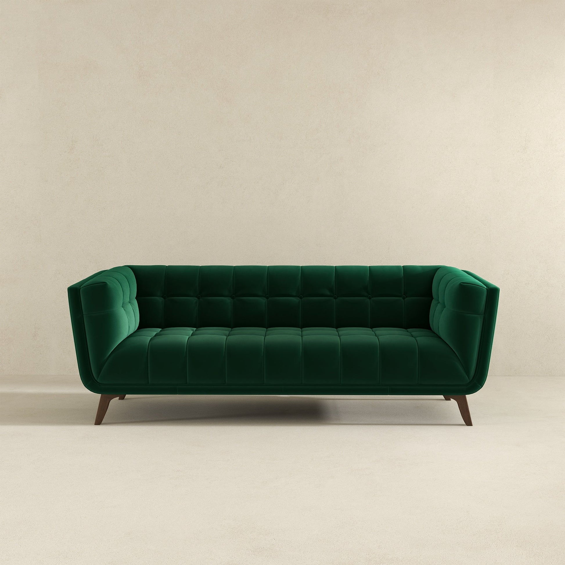 The Addison Mid-Century Modern Tufted Sofa in emerald green velvet, featuring angled wooden legs, is set against a simple beige wall and floor.