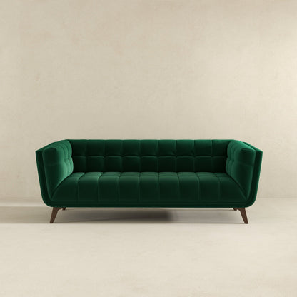 The Addison Mid-Century Modern Tufted Sofa in emerald green velvet, featuring angled wooden legs, is set against a simple beige wall and floor.