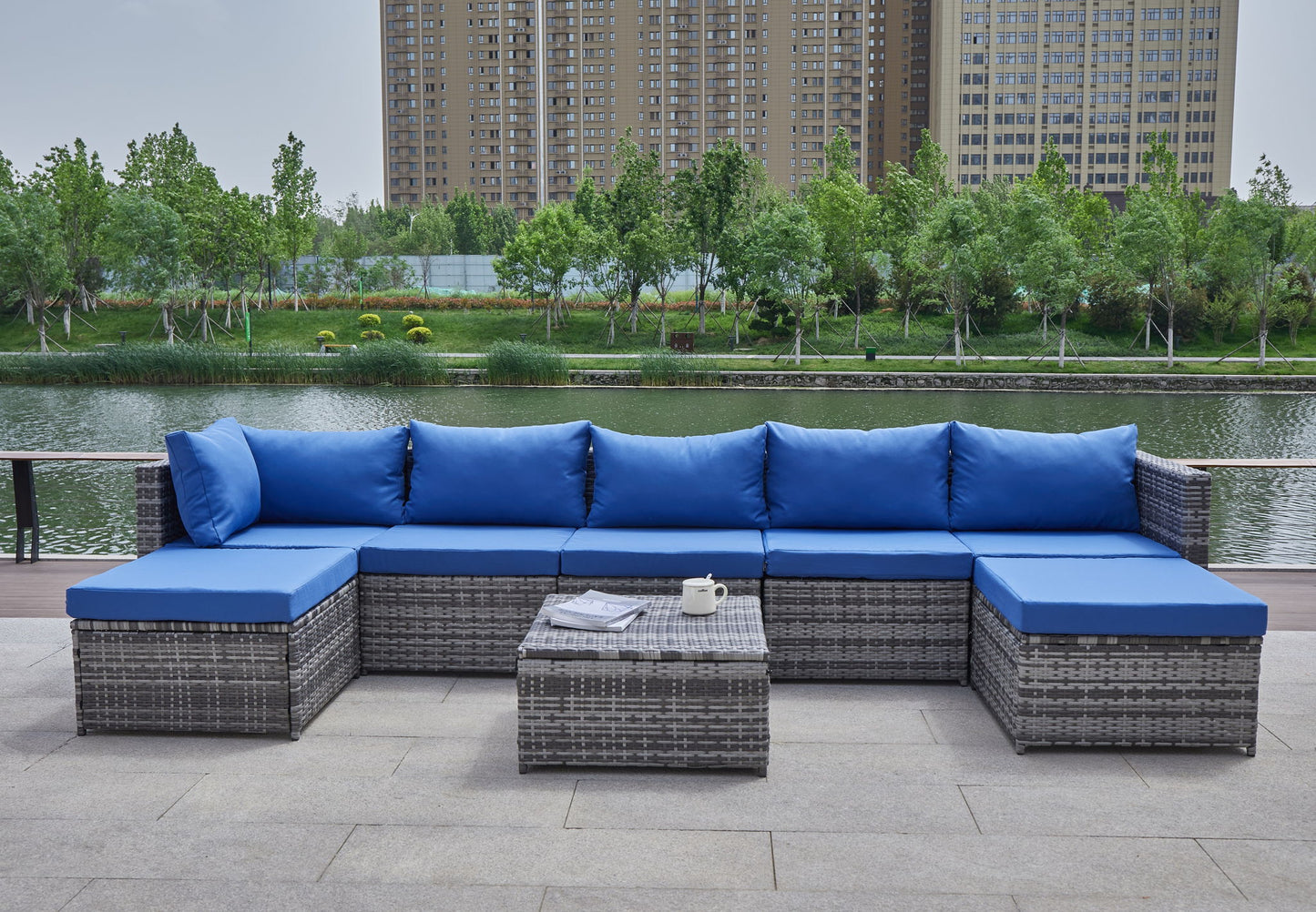 The Patio Sofa With Cushions features gray wicker sofas with bright blue cushions, a low coffee table set with a teapot and magazine, all arranged by a riverside with trees and city buildings in the background.