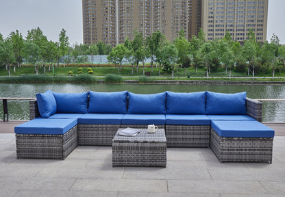 The Patio Sofa With Cushions features gray wicker sofas with bright blue cushions, a low coffee table set with a teapot and magazine, all arranged by a riverside with trees and city buildings in the background.