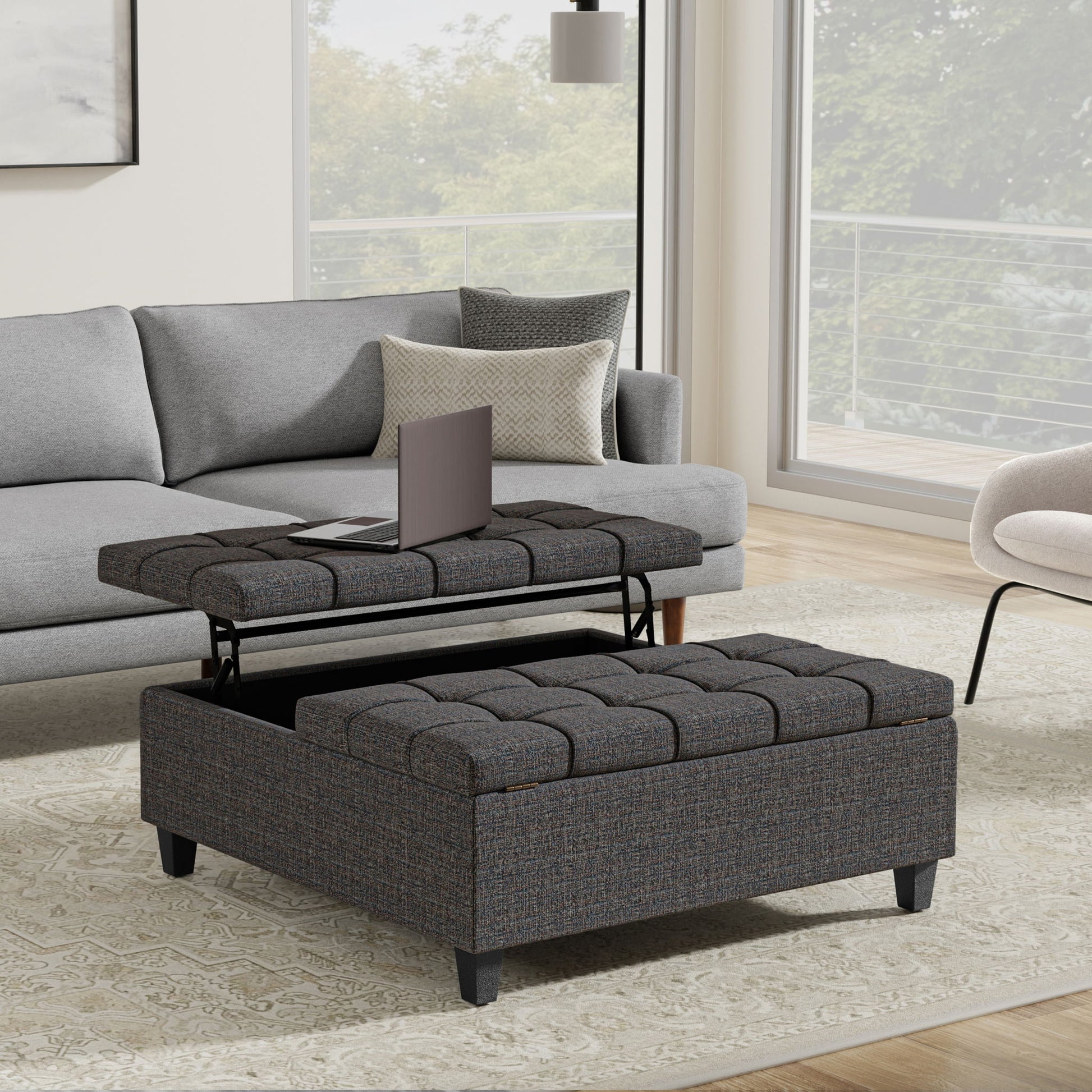 A modern living room features a gray sofa and the Harrison Upholstered Coffee Table Storage Ottoman with a lift-top for a laptop, set on a patterned rug by large windows overlooking trees and a balcony.