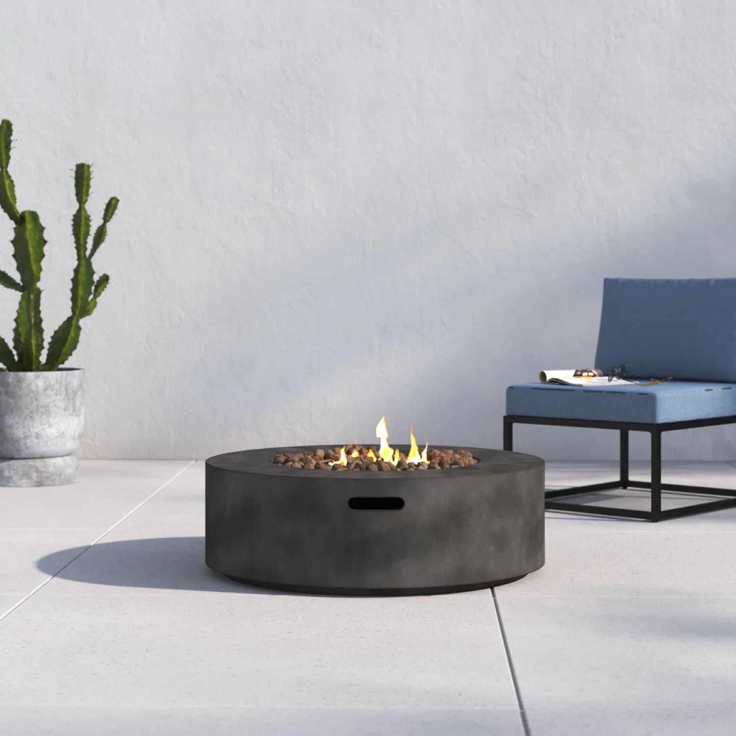 A modern patio with the Fibre Reinforced Outdoor Fire Pit Table in Charcoal burning gently, featuring a blue cushioned chair and a tall cactus in a gray pot against a light-colored wall.