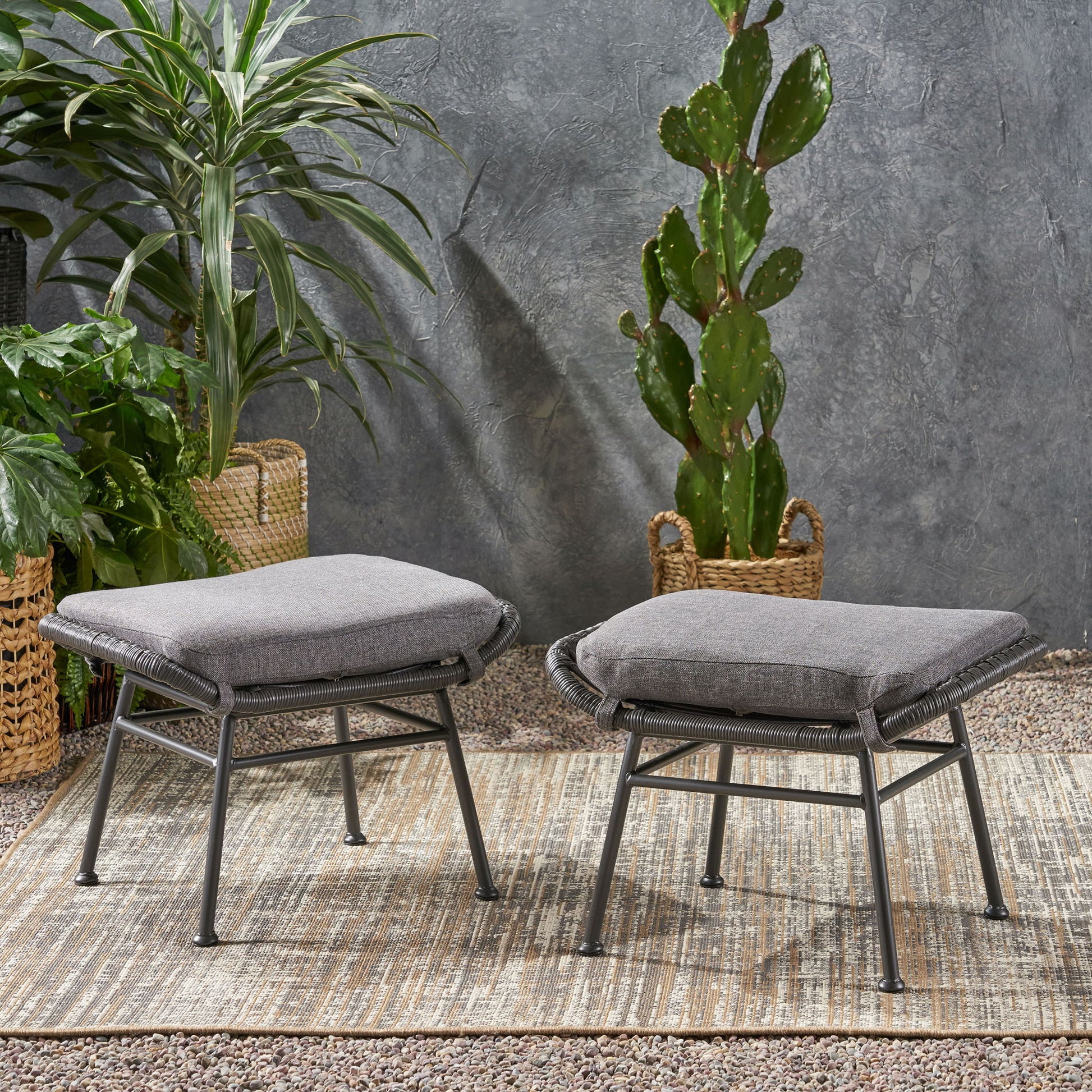 Two Outdoor Modern Boho Wicker Ottomans With Cushions (Set of 2) are arranged on a woven rug, accented by potted green plants and a textured gray wall, creating a stylish patio or garden setting.