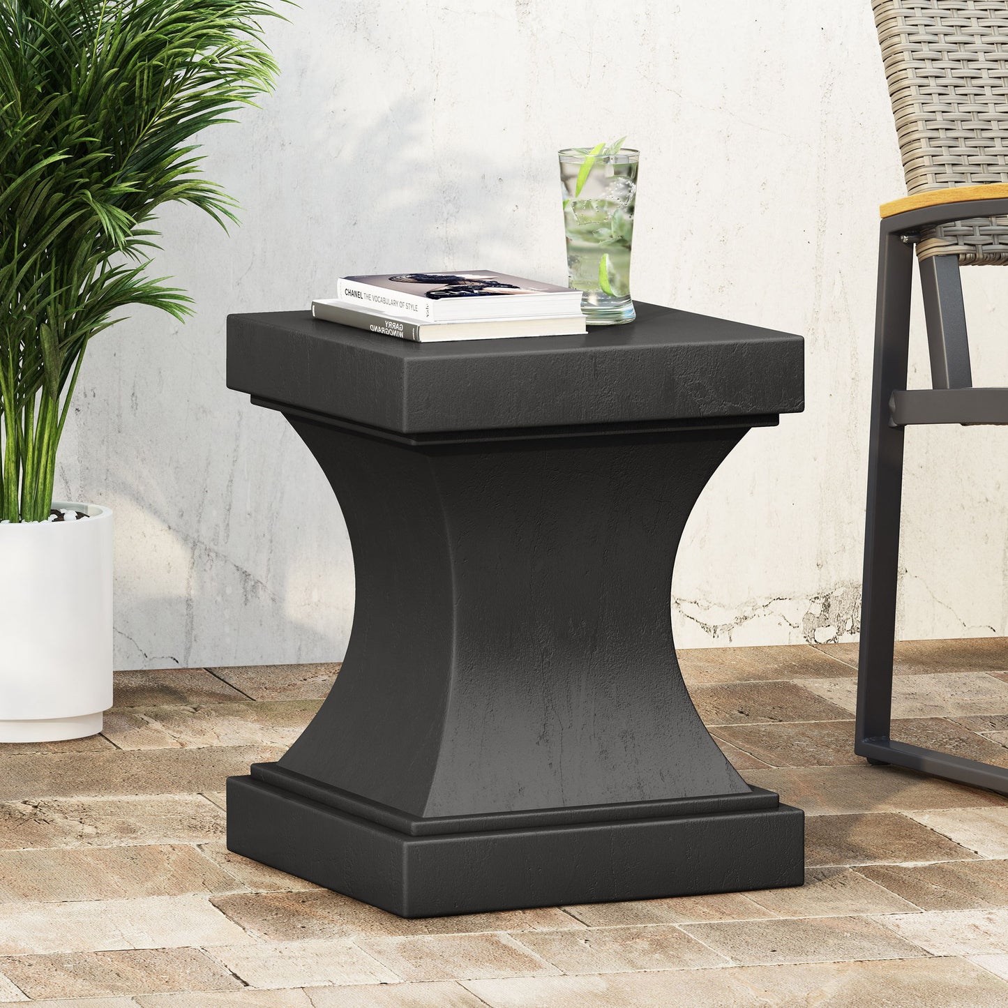 The Athena - Modern Side Table With Pedestal Design, in black, stands on a brick patio beside a chair. A glass of water and two books rest on its surface, with a potted plant nearby against a white wall.