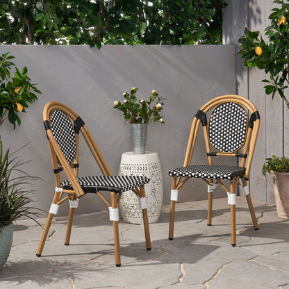 Two Outdoor Rattan And Aluminum French Bistro Chairs with black and white woven seats are set on a stone patio beside a white table with flowers, surrounded by green plants and trees, including one bearing lemons.
