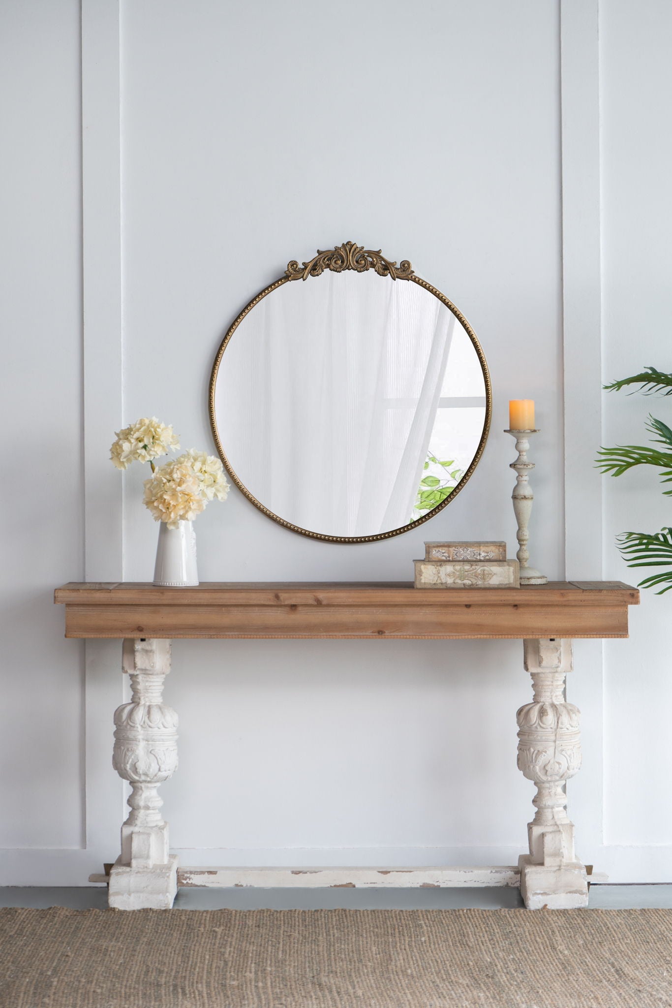 A Classic Design Mirror with a baroque-inspired frame leans above a wooden console table with ornate white legs, adorned with white flowers in a vase, two books, a lit candlestick, and a side plant.