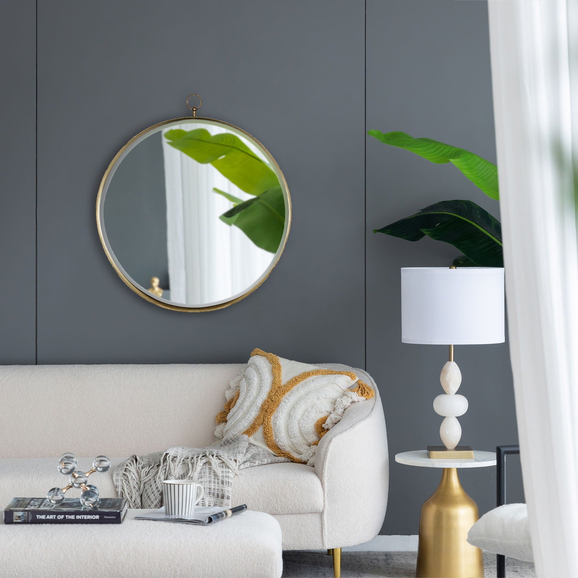 A modern living room with a Round Mirror, Circle Mirror With Iron Frame For Living Room Bedroom Vanity Entryway, Hallway reflecting green leaves; a white sofa with textured pillows, gold side table, white lamp, gray walls, and sheer curtain.