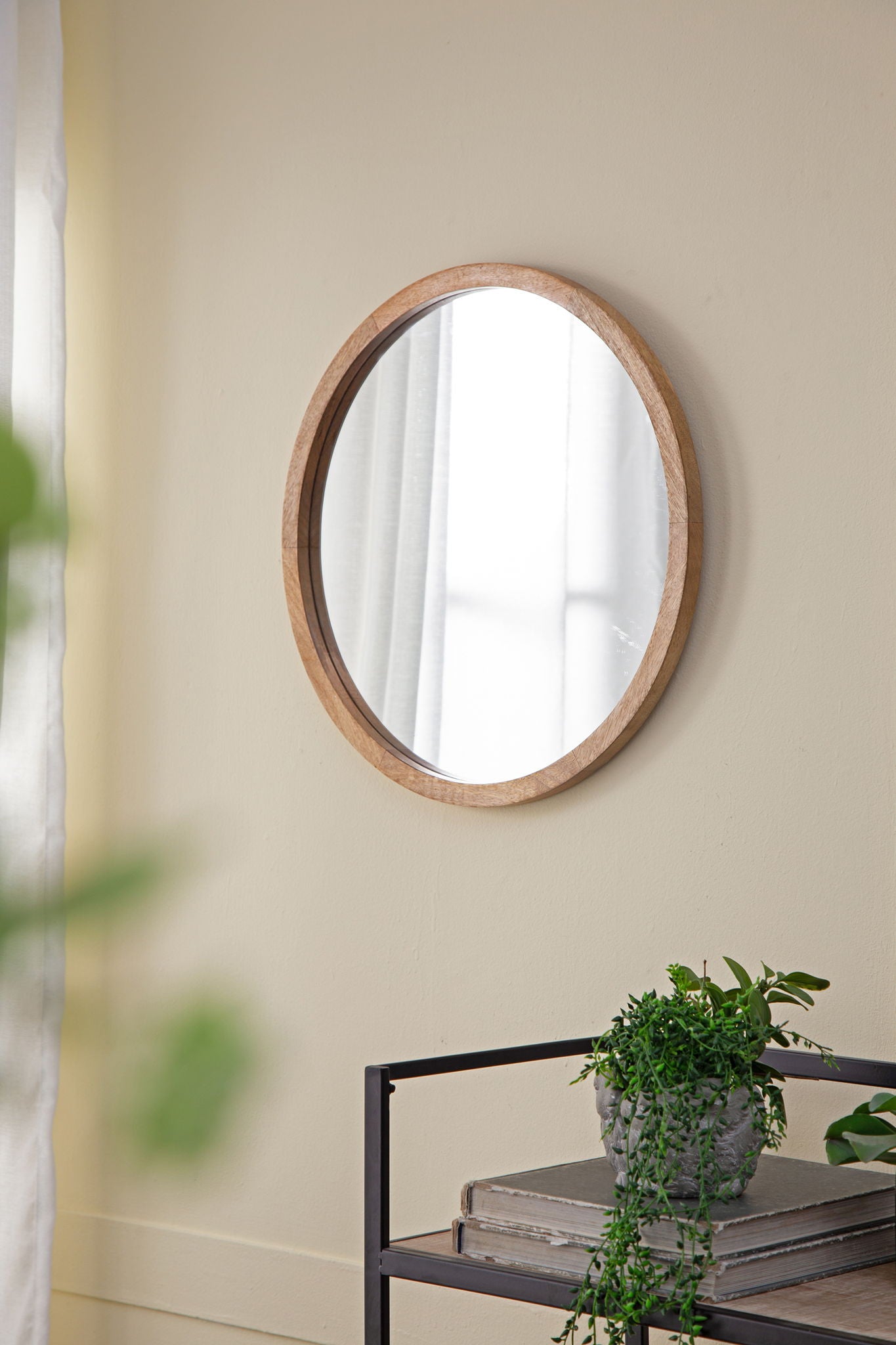 A Circle Wall Mirror With Wooden Frame hangs on a beige wall near a window with sheer curtains. Below, a black metal shelf displays books and a small potted plant with trailing green leaves.