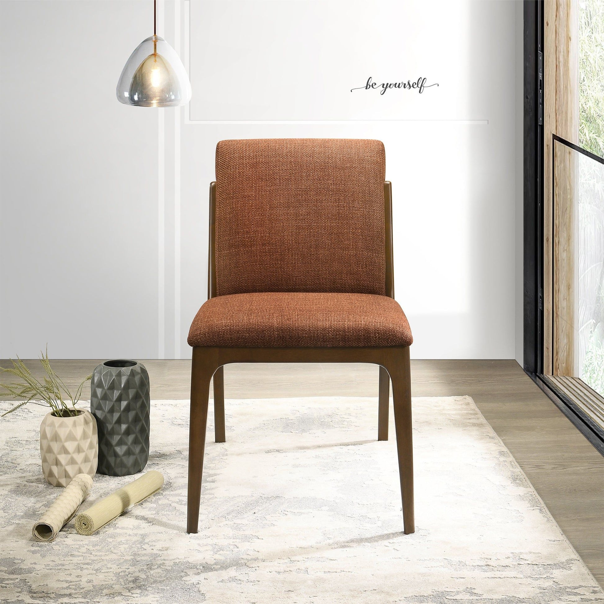 A set of Grivelle Elegant Dining Chairs in brown upholstery sits on a patterned rug in a bright room with wood flooring, decorative vases, a pendant light, and a large window. "Be yourself" is written on the wall.