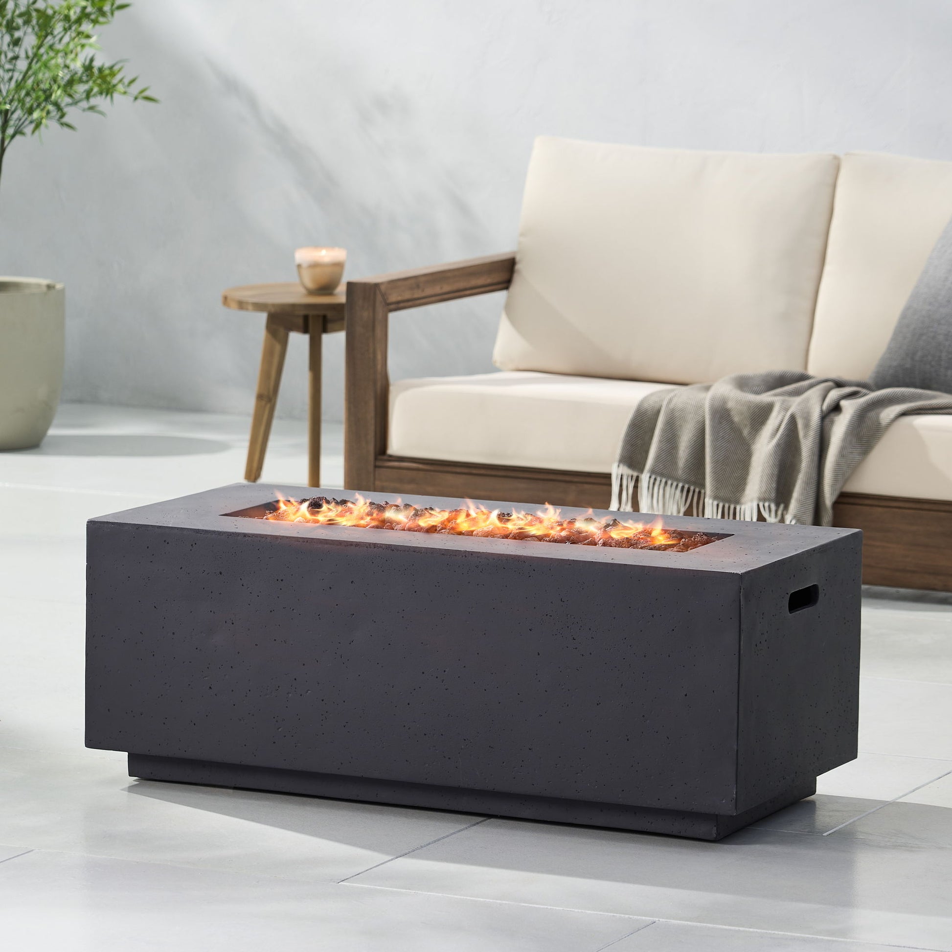 A modern patio showcases a Contemporary Outdoor 40,000 Btu Propane Fire Pit with flames, a beige cushioned wooden sofa, gray throw blanket, potted plant, and a small candle on the side table.