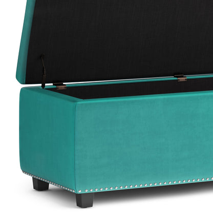 The Hamilton Upholstered Storage Ottoman in teal features a hinged lid revealing interior storage, silver nailhead trim along the bottom edge, and sturdy black wooden legs.
