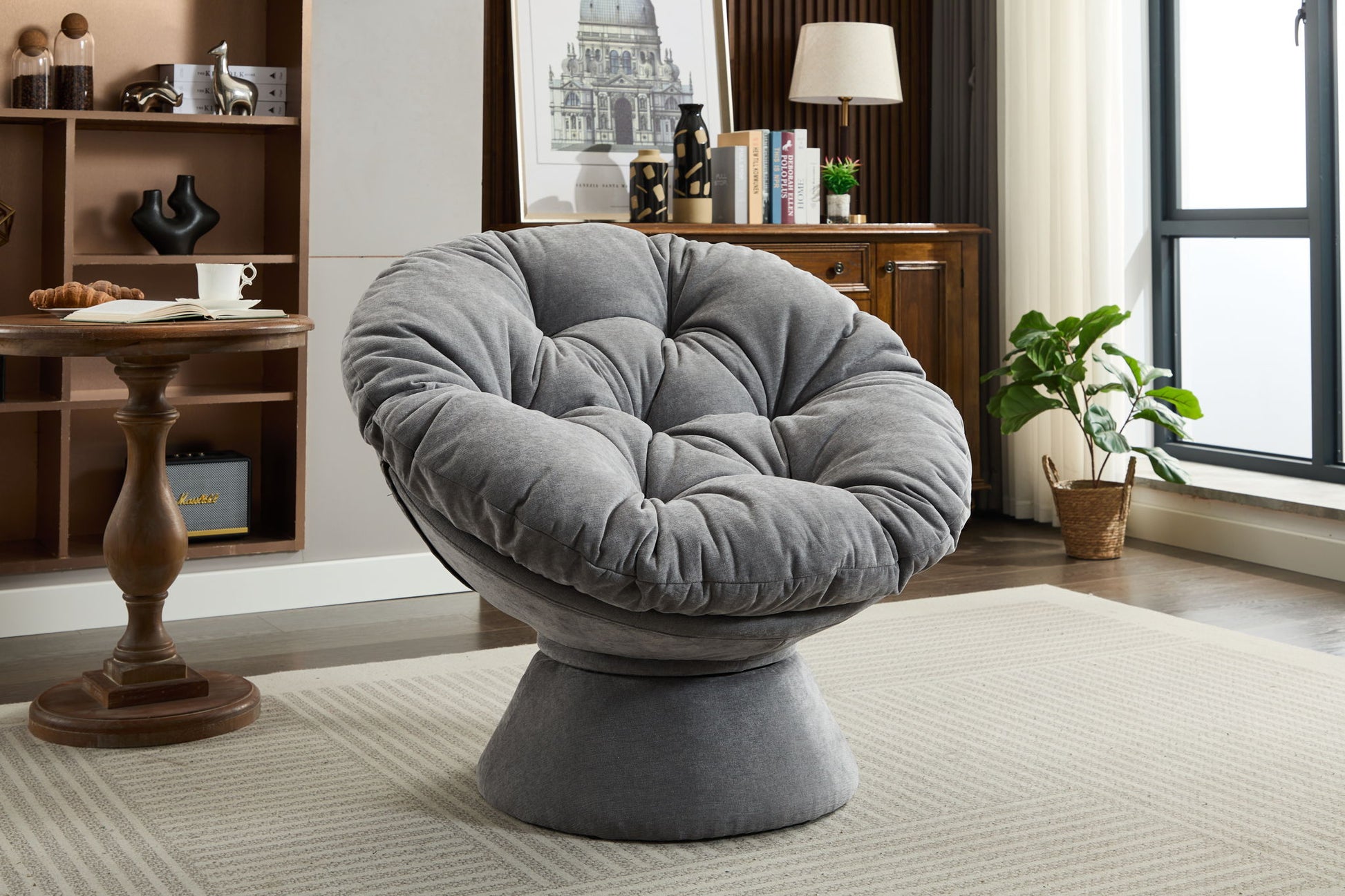 A modern living room showcases the Oversized Swivel Accent Chair, a plush, gray 360° swivel barrel papasan chair on a light rug, paired with a wooden table, cabinet, potted plant, and large window in the background.