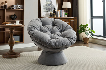 A modern living room showcases the Oversized Swivel Accent Chair, a plush, gray 360° swivel barrel papasan chair on a light rug, paired with a wooden table, cabinet, potted plant, and large window in the background.