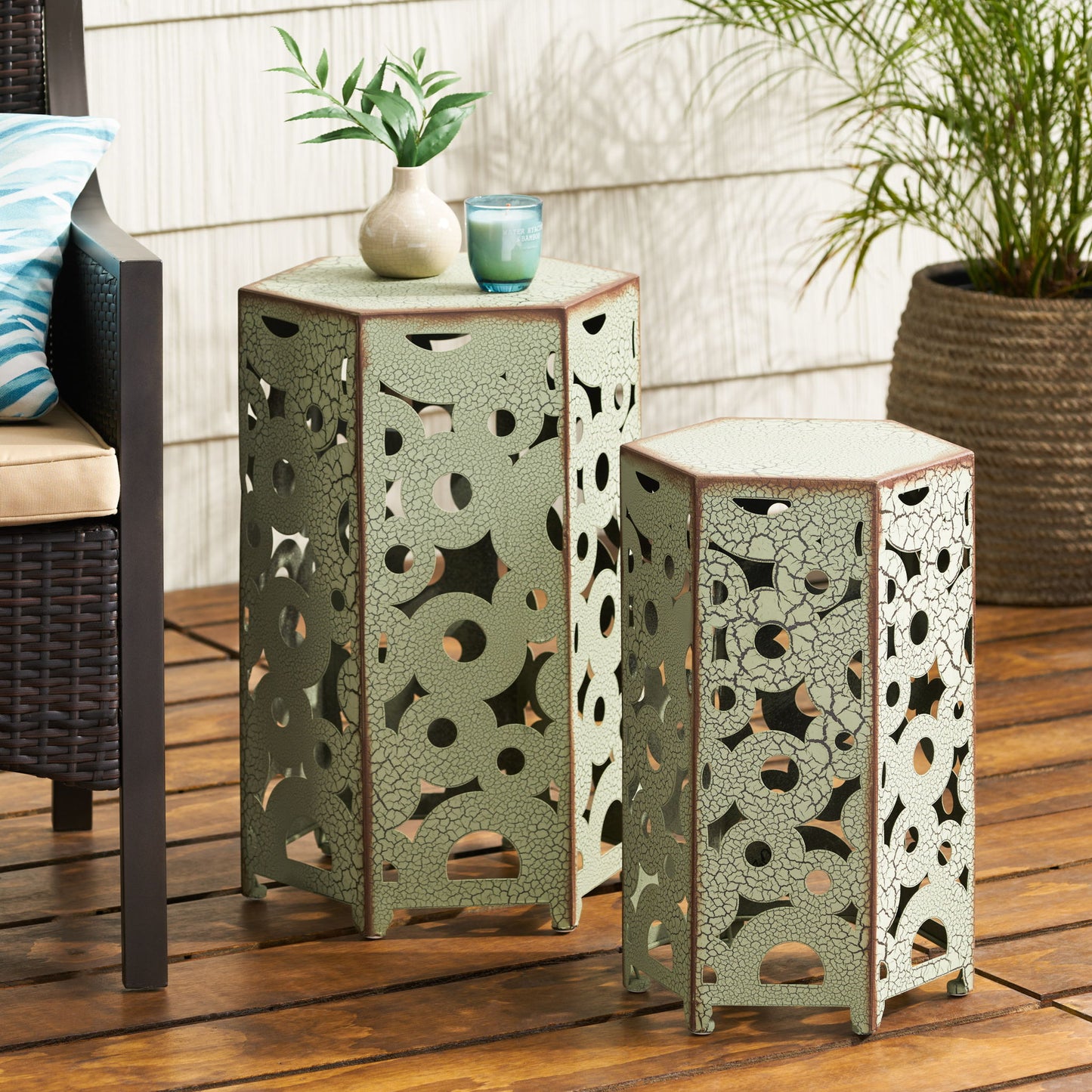 The Selva - Accent Table Set, featuring two decorative hexagonal metal tables with intricate cut-out patterns, sits on a wooden patio. A plant, candle, and cup rest on the taller table, with outdoor furniture and a potted plant nearby.