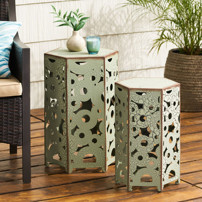The Selva - Accent Table Set, featuring two decorative hexagonal metal tables with intricate cut-out patterns, sits on a wooden patio. A plant, candle, and cup rest on the taller table, with outdoor furniture and a potted plant nearby.
