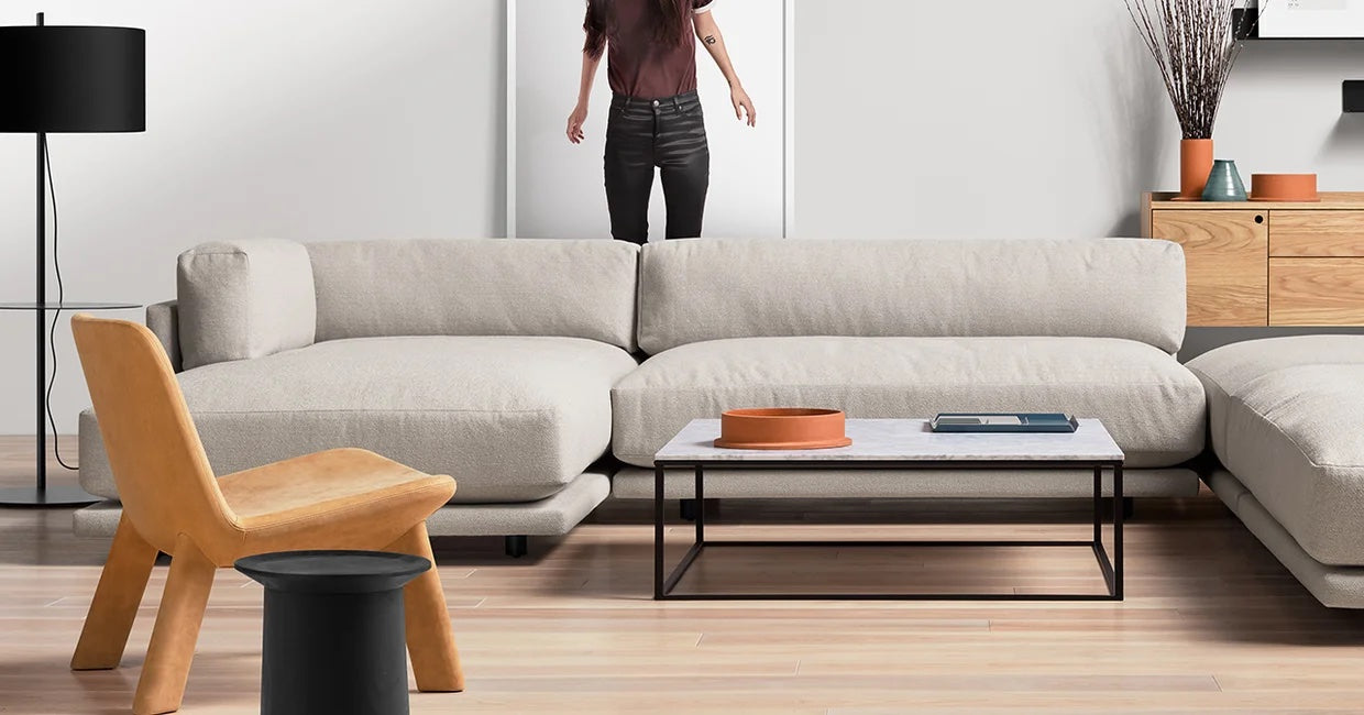 A modern living room featuring a large beige sectional sofa, a light wooden floor, a black floor lamp, and a low coffee table with an orange tray and a book. A mustard-colored chair and a black side table are also present. A person stands in the background.