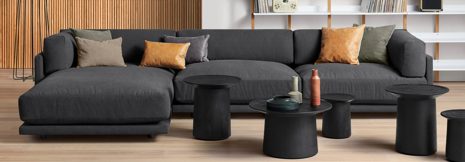 A modern living room features a dark gray sectional sofa adorned with various colored pillows, including brown, tan, and green. In front of the sofa are three black round coffee tables with decorative items. A wooden slat wall and shelves with books and records are in the background.