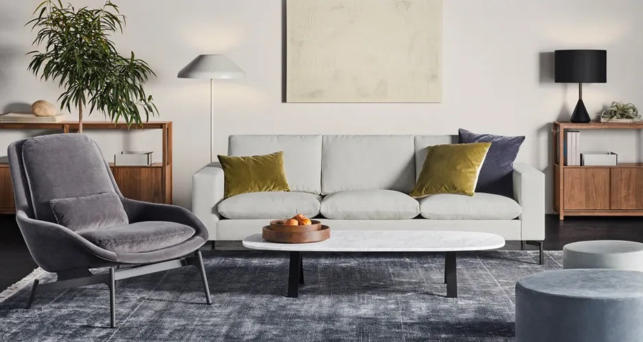 A modern living room features a large gray sectional sofa with several dark gray and light gray cushions, a white marble coffee table with pink flowers in a vase, a round gray ottoman, and a floor lamp. The furniture sits on a dark textured rug.