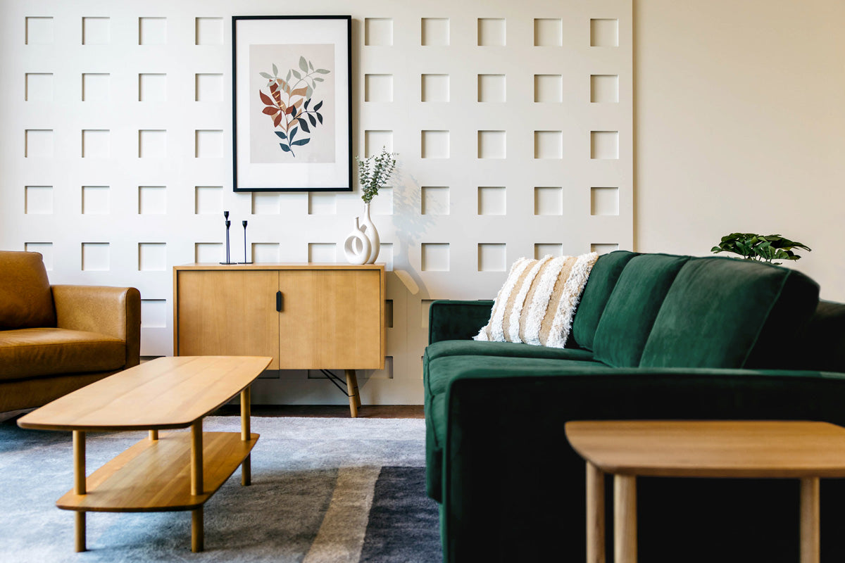 A modern living room with a green velvet sofa, a wooden coffee table, and a brown armchair. A wooden sideboard with decorative vases and a framed abstract leaf print hang on a textured white wall. A rug covers part of the floor.