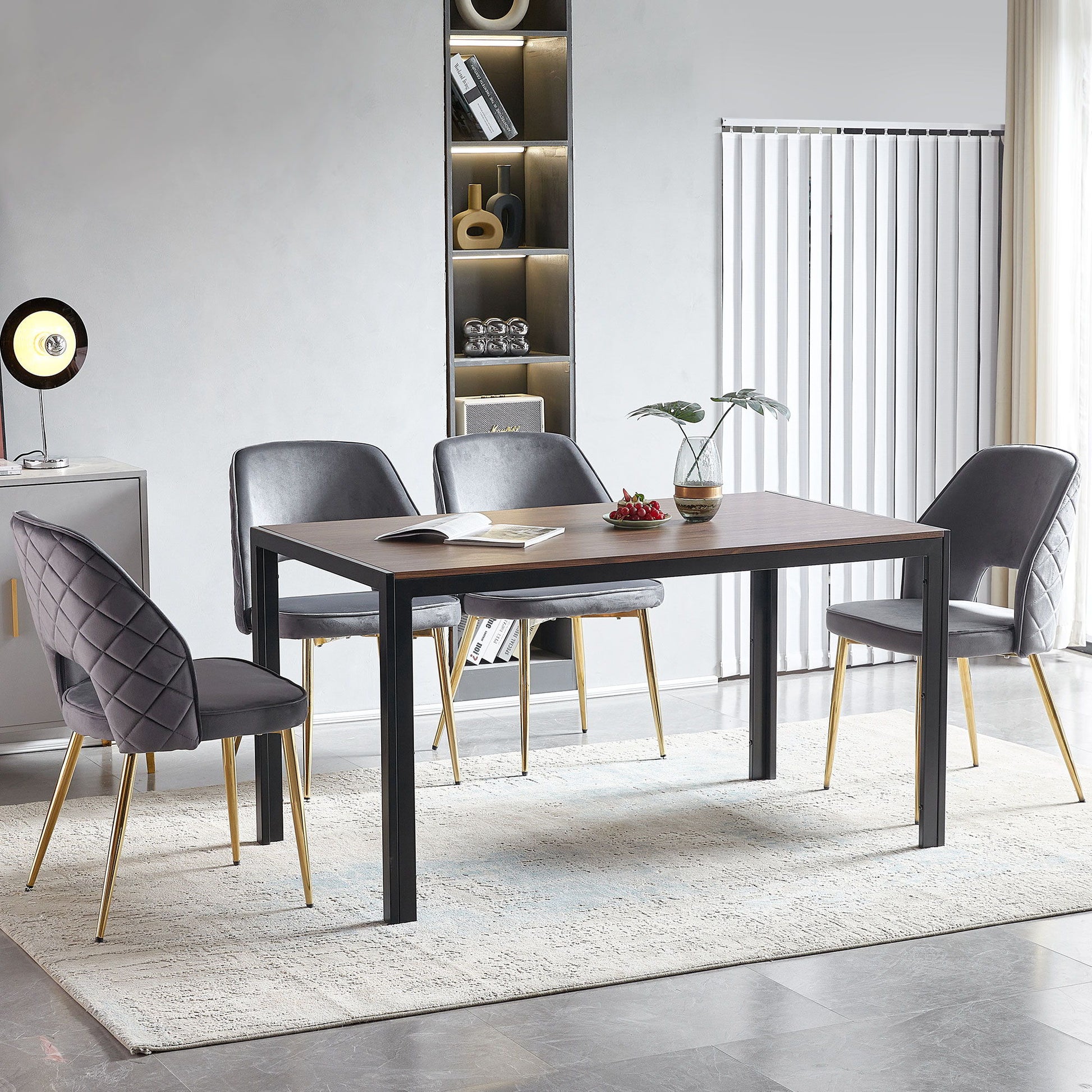 A modern dining room features a rectangular wooden table with four Velvet Dining Chairs With Metal Legs And Hollow Back Upholstered Dining Chairs on a light rug. A small plant and books are on the table, with built-in shelves and vertical blinds in the background.