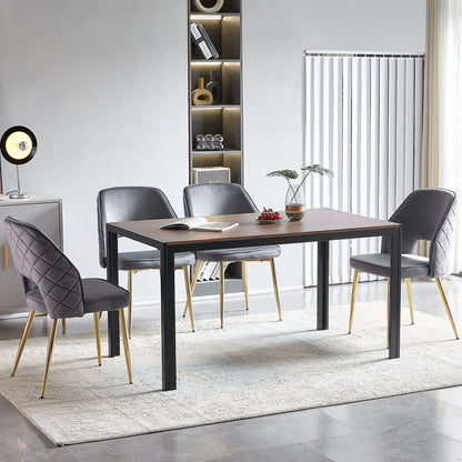 A modern dining room features a rectangular wooden table with four Velvet Dining Chairs With Metal Legs And Hollow Back Upholstered Dining Chairs on a light rug. A small plant and books are on the table, with built-in shelves and vertical blinds in the background.