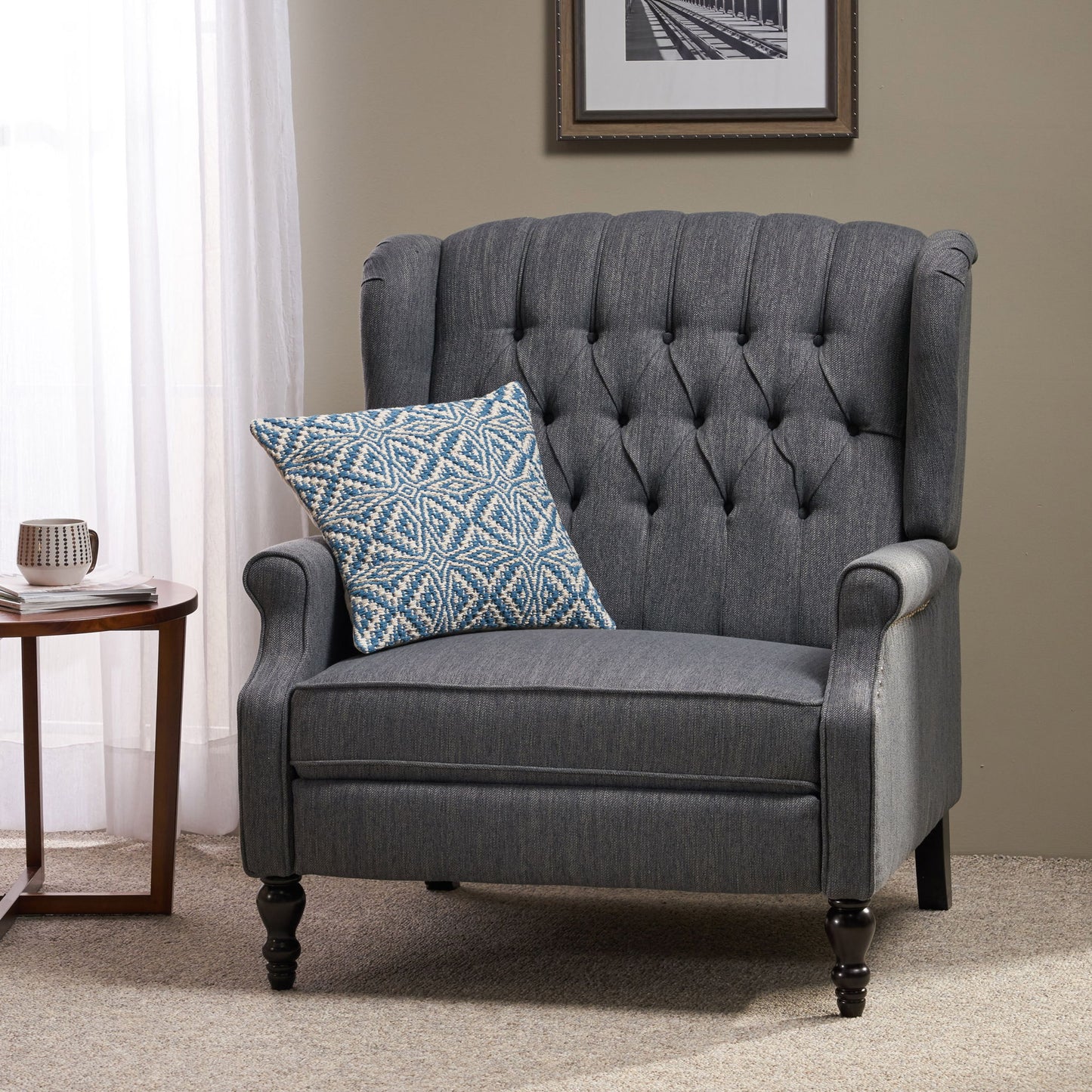 A Dunham Contemporary Fabric Tufted Oversized Pushback Recliner with a blue and white pillow sits on beige carpet beside a small round table holding a mug and book, near a window with white curtains and a framed photo.