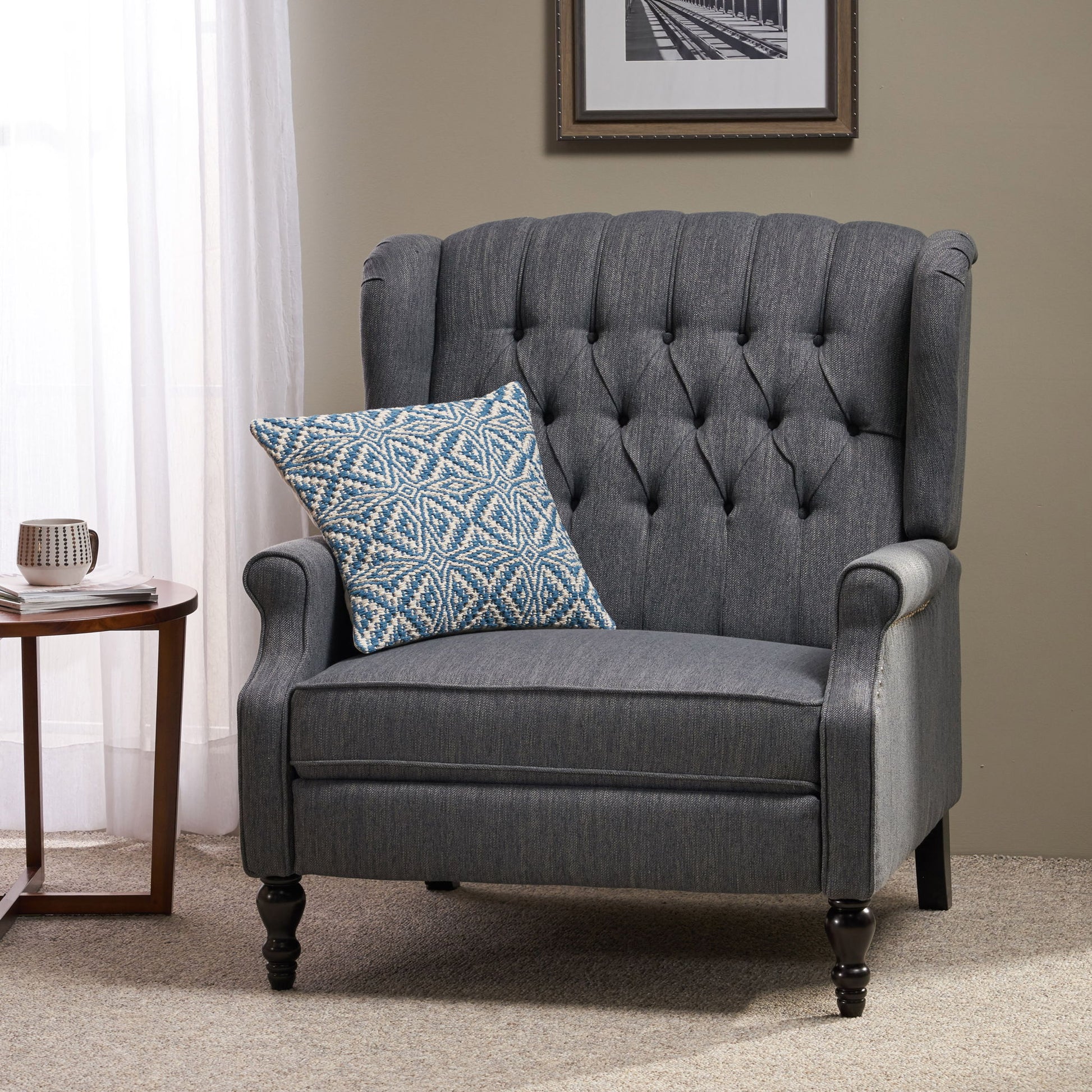 A Dunham Contemporary Fabric Tufted Oversized Pushback Recliner with a blue and white pillow sits on beige carpet beside a small round table holding a mug and book, near a window with white curtains and a framed photo.