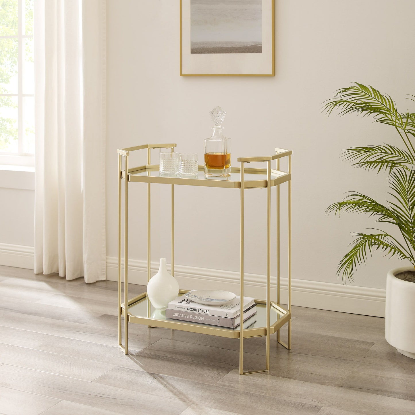 A modern pale gold accent table with a mirror top and metal construction holds a decanter, glasses, books, and a white vase. It's placed on a light wood floor near a window with white curtains, potted plant, and framed wall art.