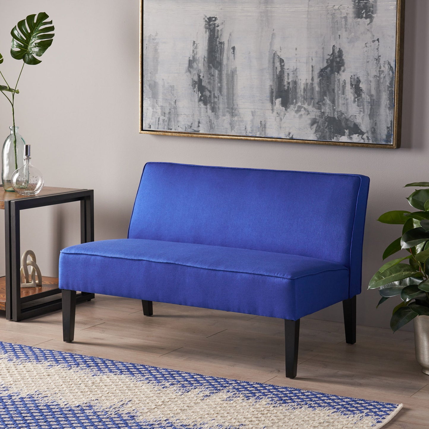 A modern room featuring the Contemporary Loveseat with upholstered fabric and birch wood legs, accented by a large abstract gray painting, green plants, a glass vase on a black side table, and a blue-and-white patterned rug.