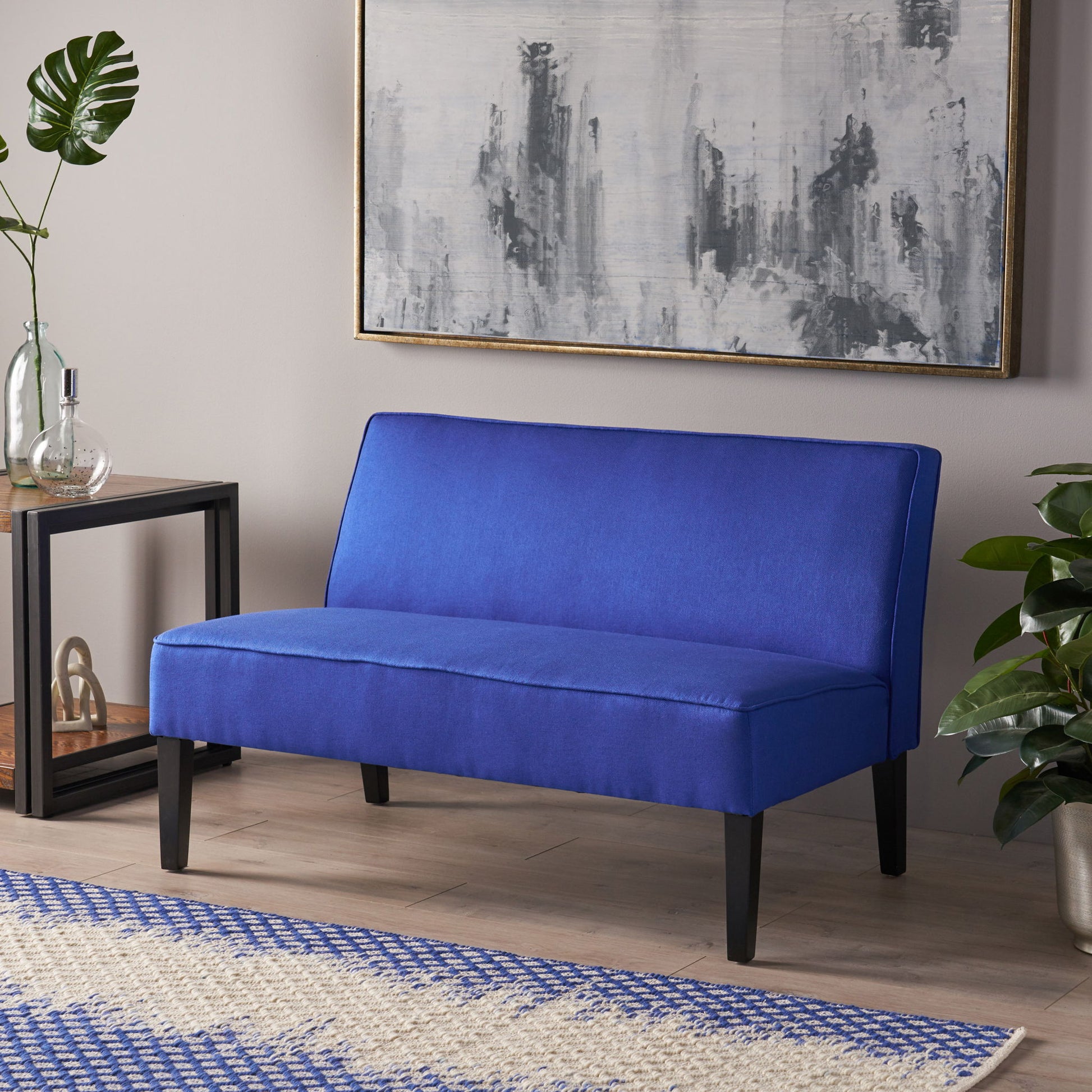 A modern room featuring the Contemporary Loveseat with upholstered fabric and birch wood legs, accented by a large abstract gray painting, green plants, a glass vase on a black side table, and a blue-and-white patterned rug.