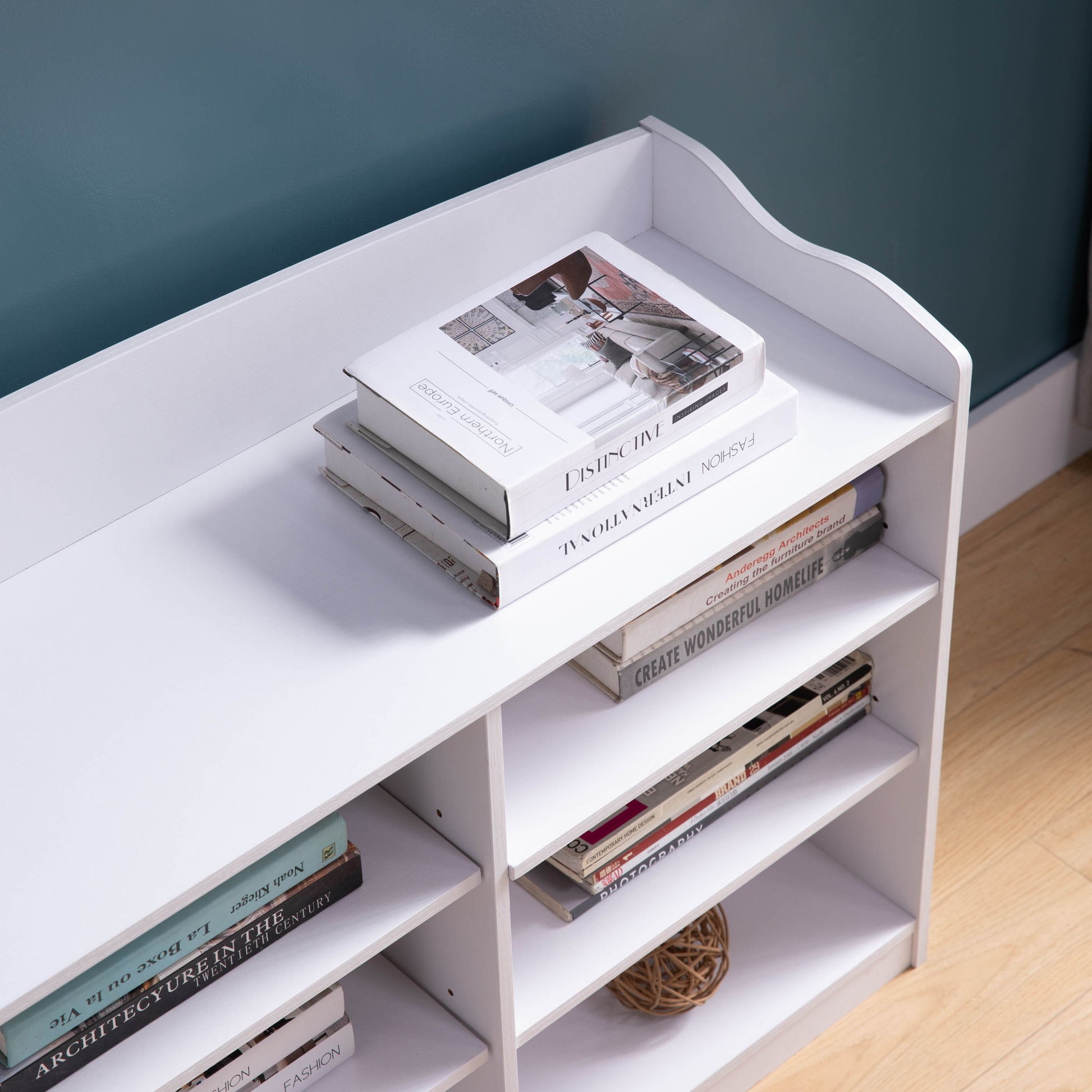 A white Entryway Shoe Storage Bench with nine shelves sits against a teal wall, holding stacked books and magazines on top, plus more books and a decorative rattan ball below. Light wood flooring is partially visible.
