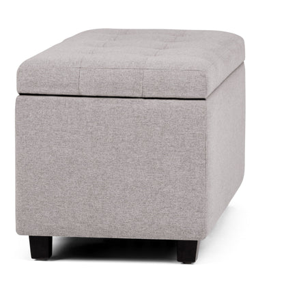 The Cosmopolitan Upholstered Storage Ottoman features light gray fabric, a padded tufted lid, short black legs, and a hollow storage space visible with the lid slightly open.