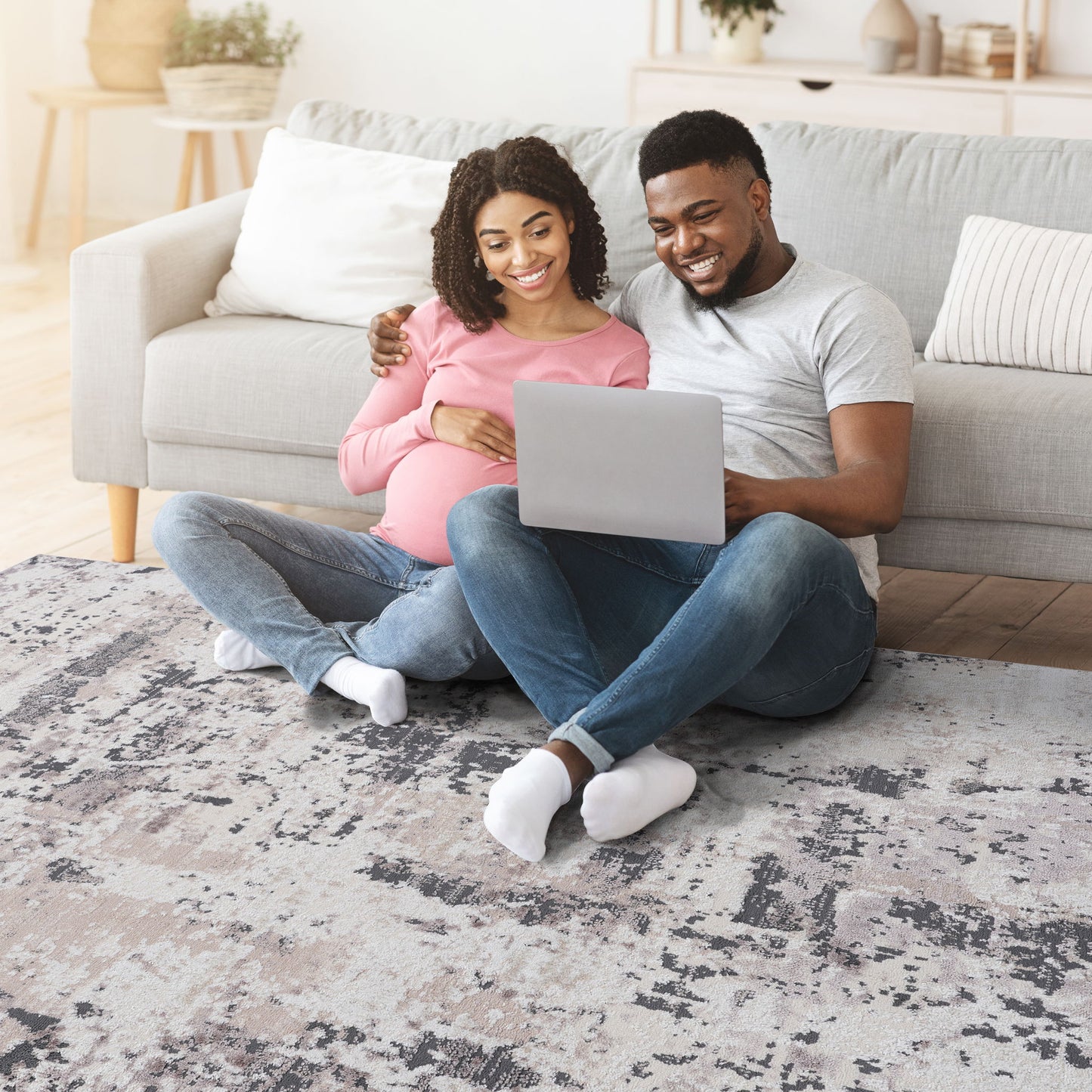 A smiling couple sits on the Payas - Abstract Area Rug in front of their sofa, looking at a laptop together. The pregnant woman rests her hand on her belly as the man wraps his arm around her shoulders.