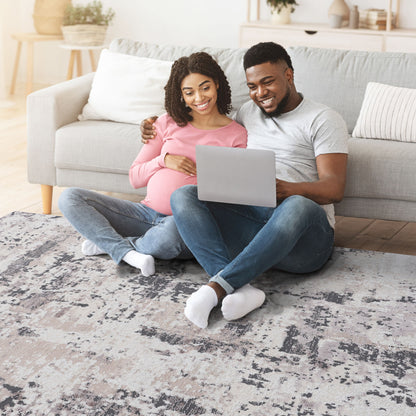 A smiling couple sits on the Payas - Abstract Area Rug in front of their sofa, looking at a laptop together. The pregnant woman rests her hand on her belly as the man wraps his arm around her shoulders.