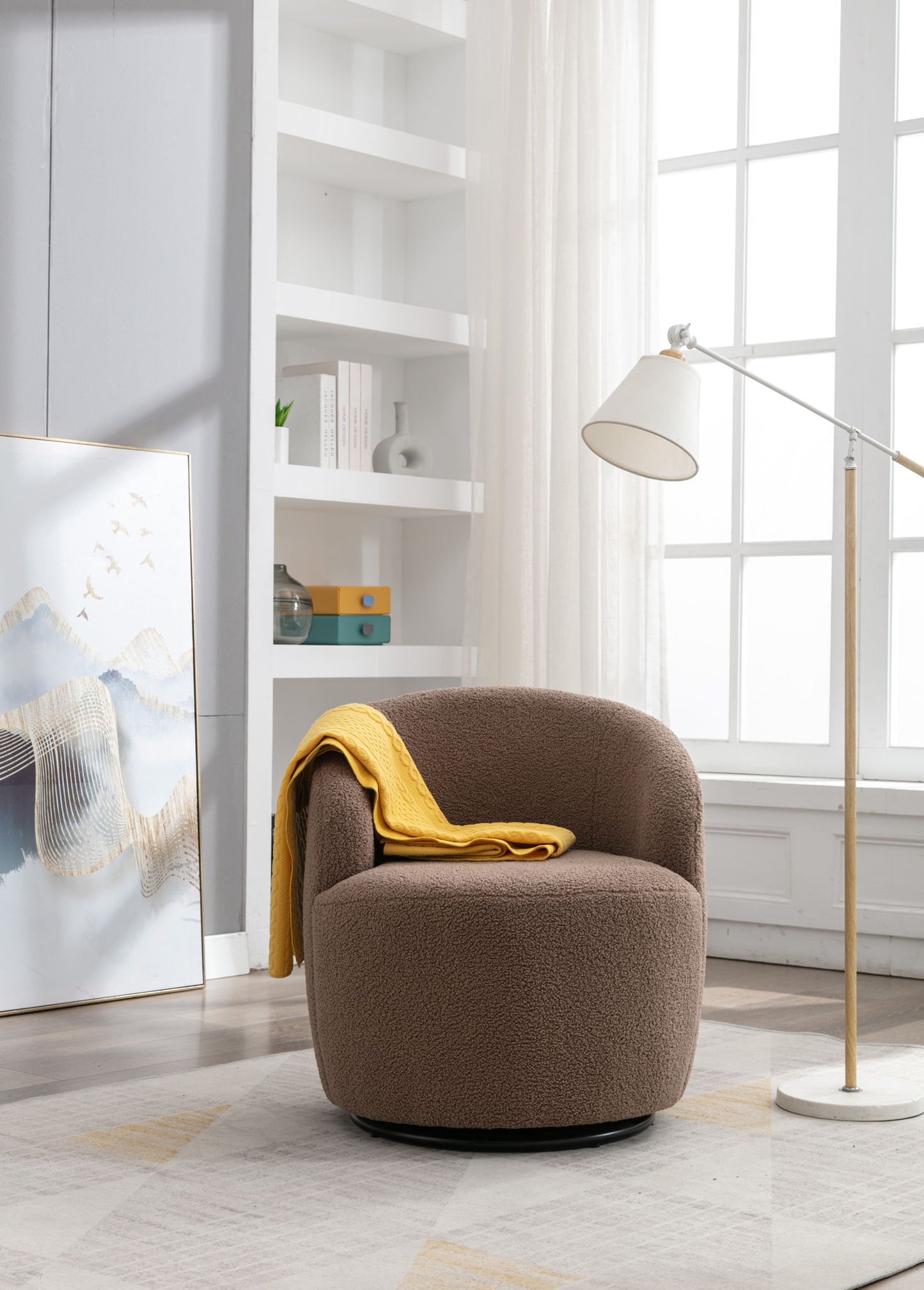 A Teddy Fabric Swivel Accent Armchair Barrel Chair with a yellow blanket sits by a tall floor lamp in a bright modern room featuring white shelves, large windows, and framed artwork on the floor.
