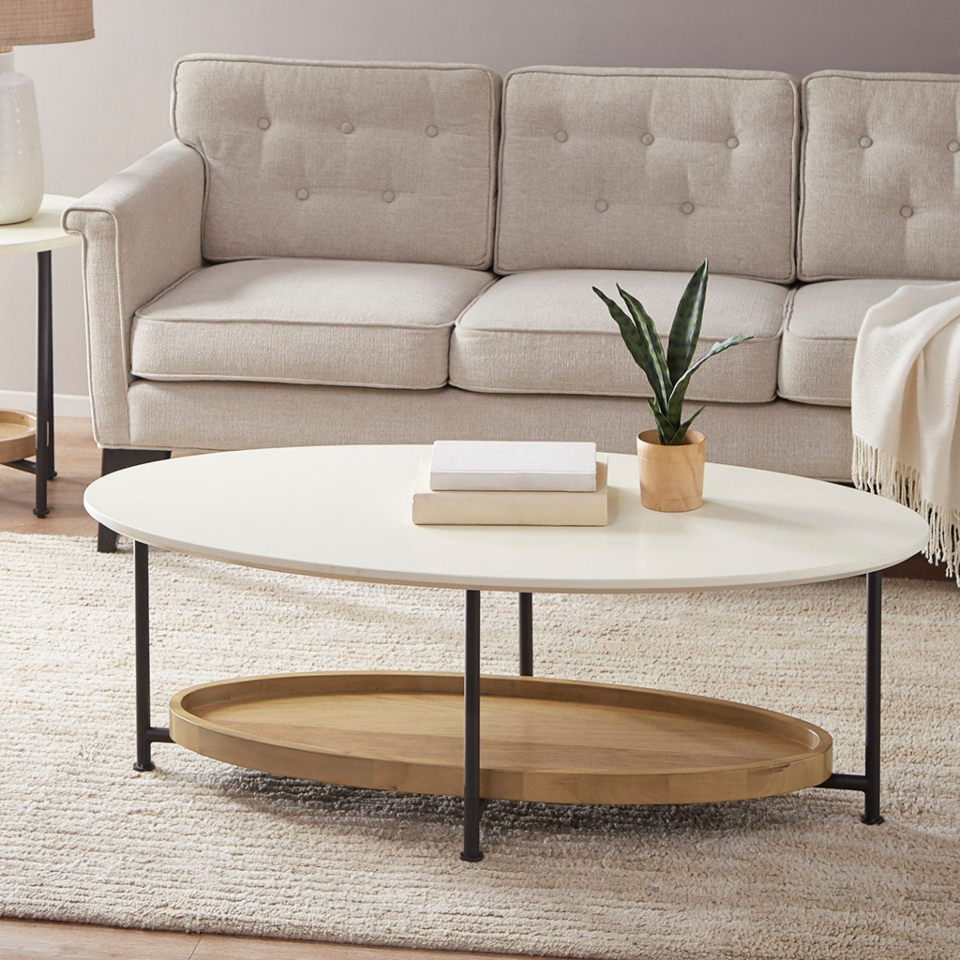 A modern living room featuring a light beige sofa, the Oval Coffee Table With Metal Legs - White / Natural with books and a potted plant on top, all set on a textured cream rug.