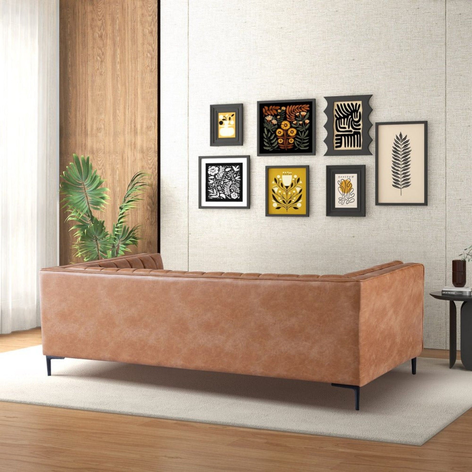 A modern living room with the Angelina - Channel Tufted Sofa in tan leather on a light rug, a tall green plant, and a wooden accent wall. Six framed botanical and abstract artworks decorate the cream-colored wall.