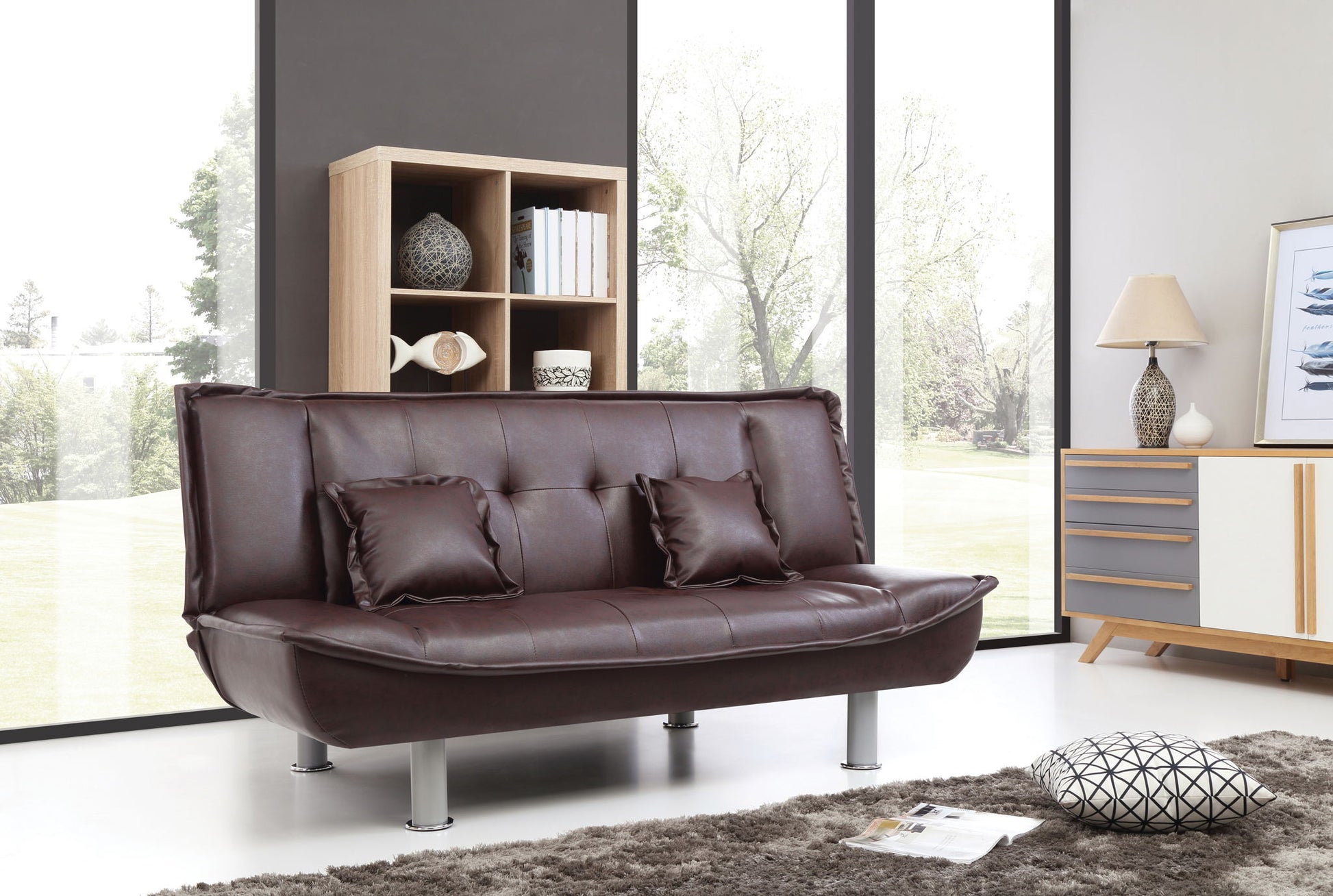A modern living room featuring the Lionel - Sofa Bed in dark brown leather, two matching cushions, a shaggy gray rug, a patterned pillow, wooden shelf, sideboard, lamp, and large windows overlooking trees.