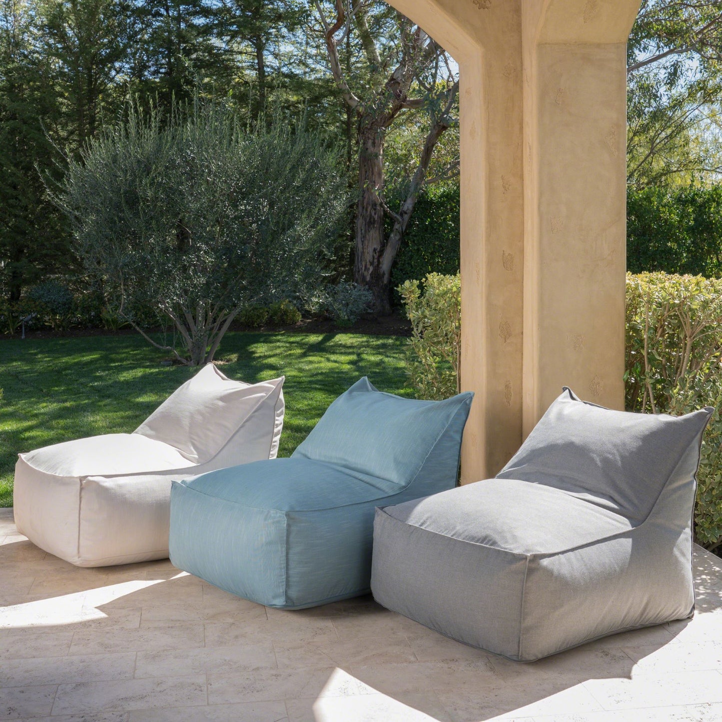 Three Ettie Water Resistant Fabric Bean Bag Chairs in white, blue, and gray are arranged side by side on a stone patio with a garden backdrop, sunlight filtering through for a relaxing outdoor setting.