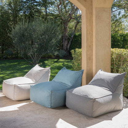 Three Ettie Water Resistant Fabric Bean Bag Chairs in white, blue, and gray are arranged side by side on a stone patio with a garden backdrop, sunlight filtering through for a relaxing outdoor setting.