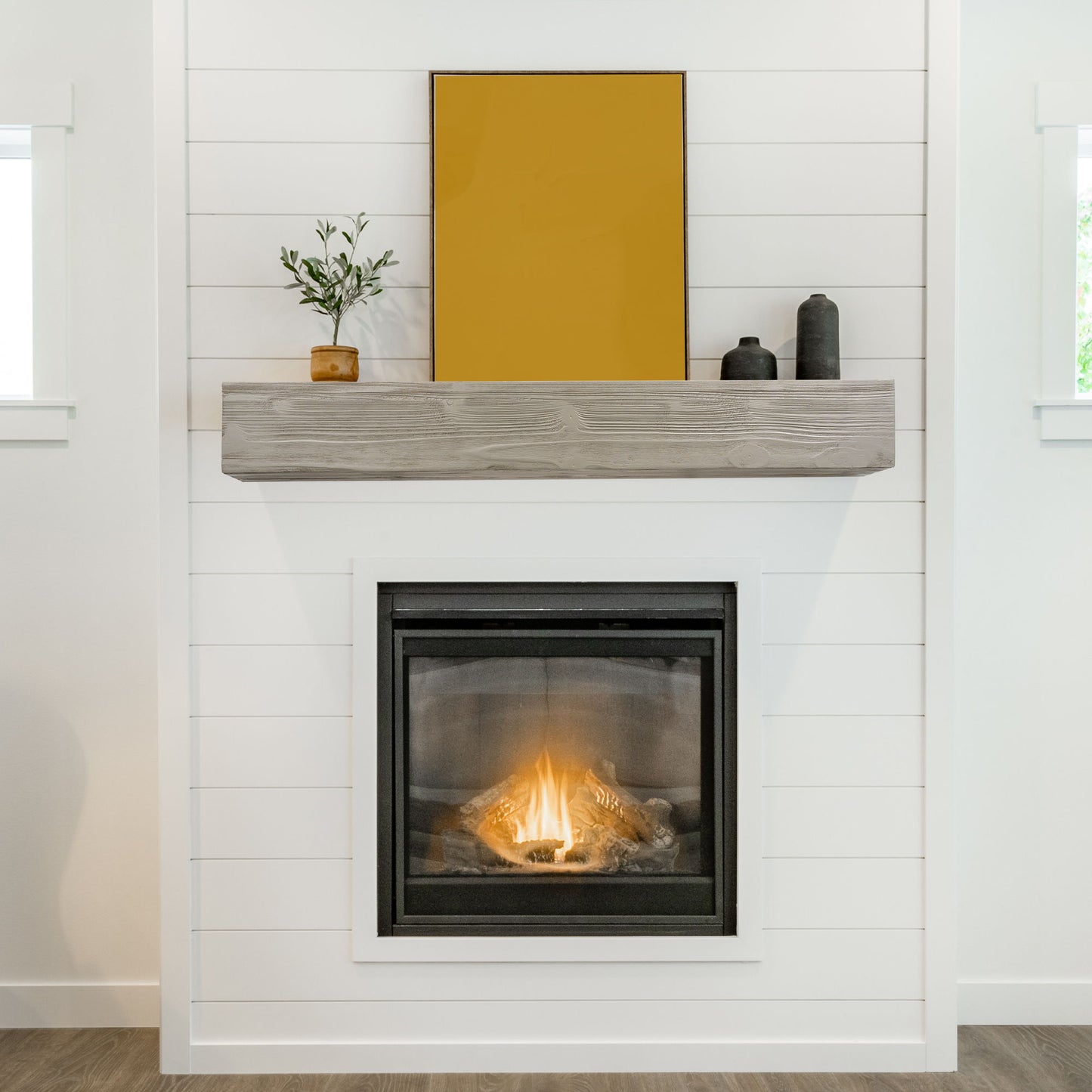 A modern white shiplap fireplace features the Rustic Wood Fireplace Mantel, wall-mounted and floating, with a potted plant, two black decorative objects, and a large yellow-framed artwork above. The lit gas fire creates a warm ambiance.