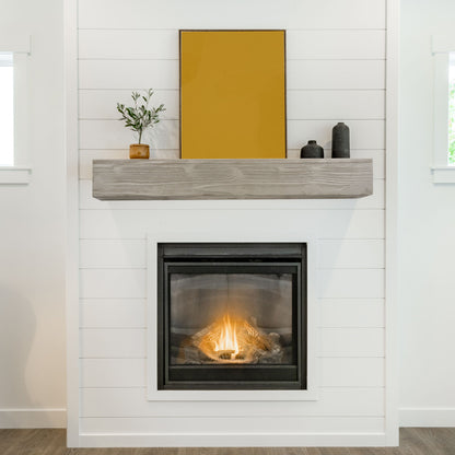 A modern white shiplap fireplace features the Rustic Wood Fireplace Mantel, wall-mounted and floating, with a potted plant, two black decorative objects, and a large yellow-framed artwork above. The lit gas fire creates a warm ambiance.