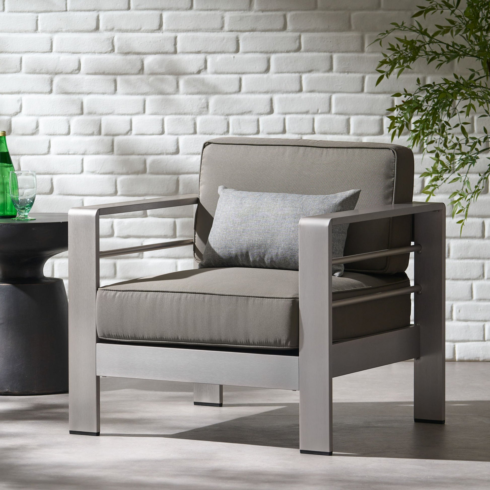 The Outdoor Aluminum Club Chair With Cushion - Gray sits on a light floor near a black side table with a green bottle. A white brick wall and leafy plant are in the background.