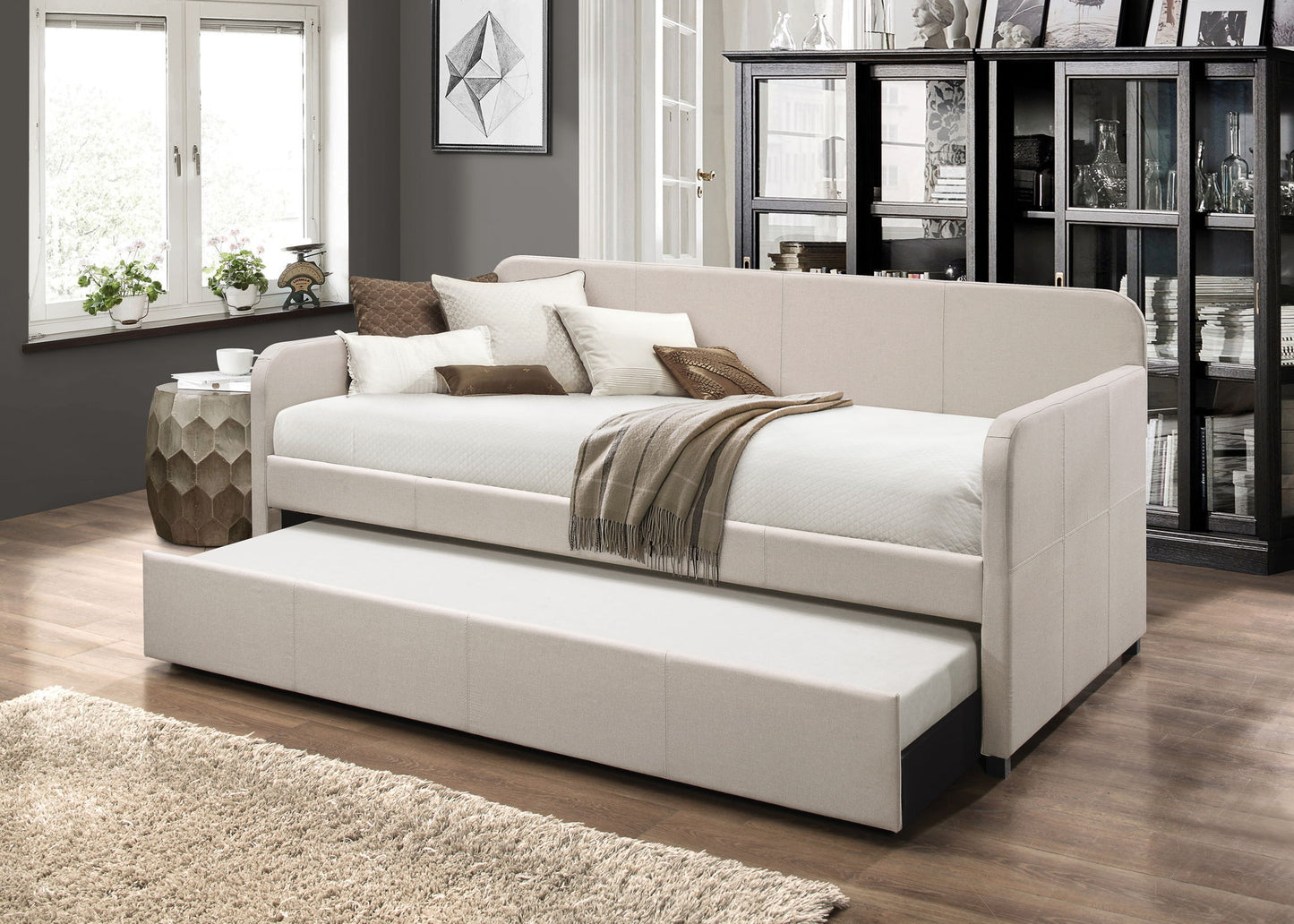 A modern living room features the Jagger Twin Daybed with Trundle in Fog, styled with cushions and a brown throw. A large window, glass-fronted cabinet, and neutral decor complete the cozy, elegant space.
