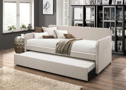 A modern living room features the Jagger Twin Daybed with Trundle in Fog, styled with cushions and a brown throw. A large window, glass-fronted cabinet, and neutral decor complete the cozy, elegant space.