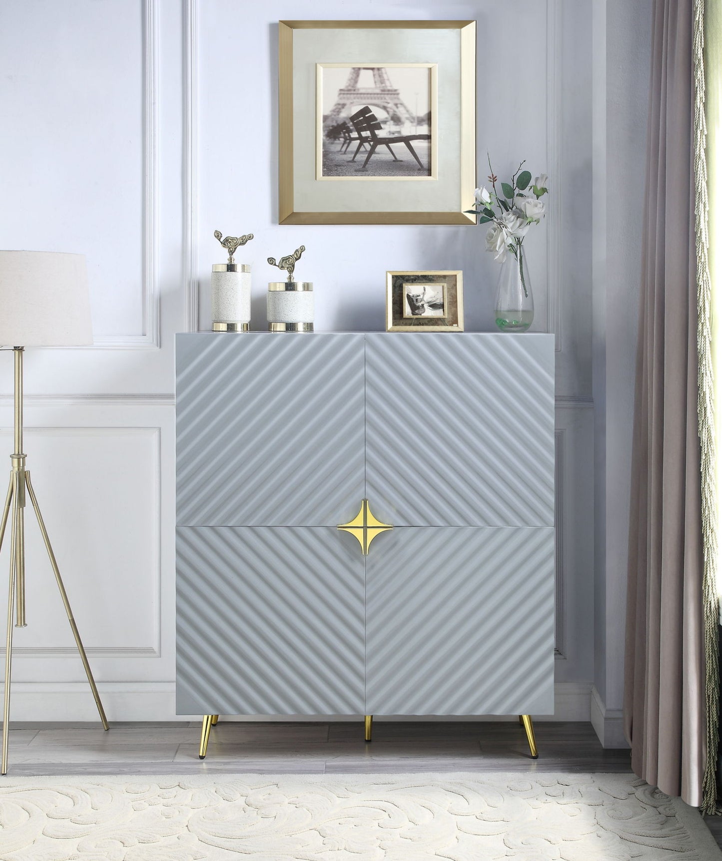 A modern room featuring the Gaines High Gloss Accent Cabinet in gray with gold accents and gold legs. Decorative items, a flower vase, and framed photos sit on top. A floor lamp and curtains flank each side.