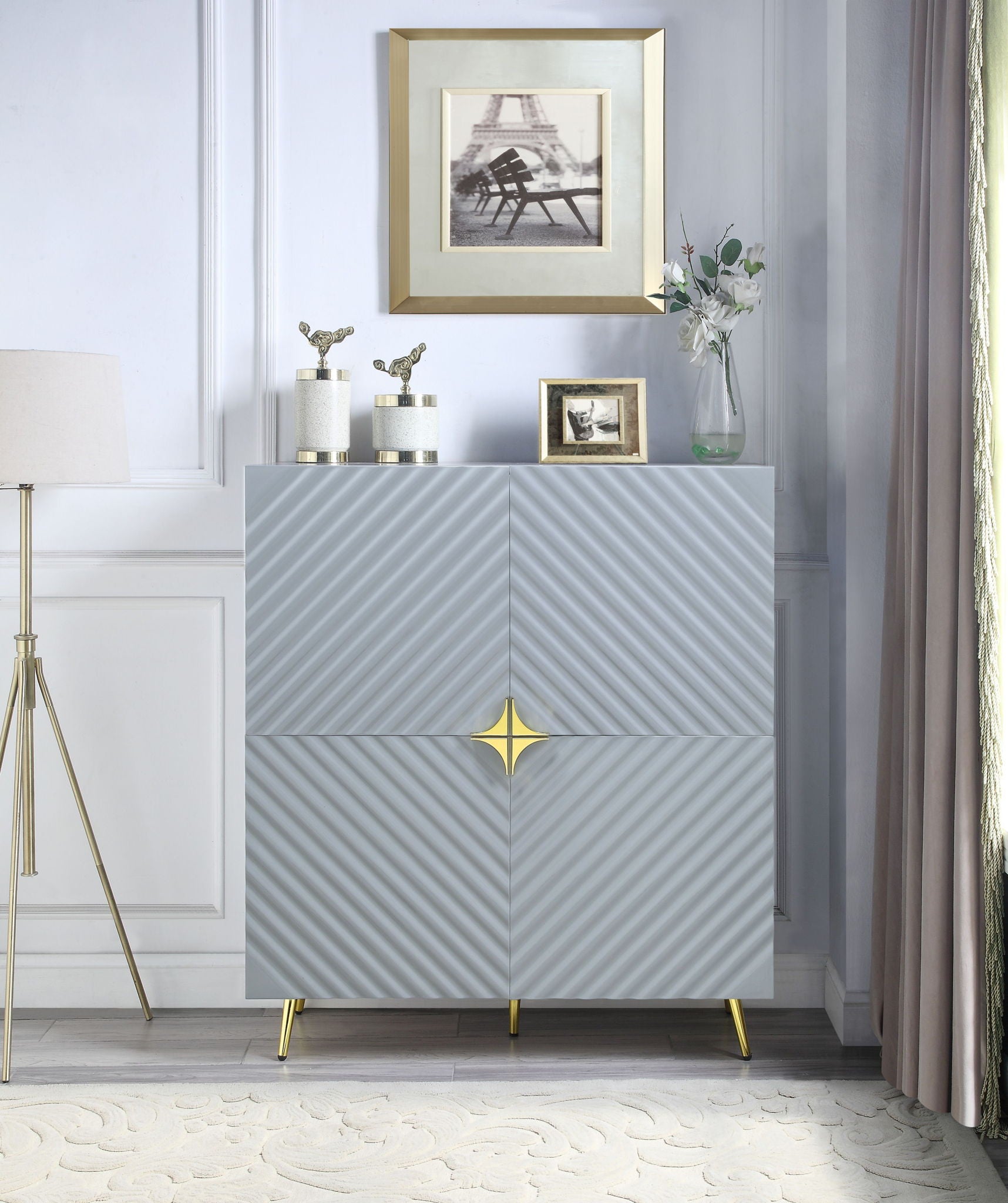 A modern room featuring the Gaines High Gloss Accent Cabinet in gray with gold accents and gold legs. Decorative items, a flower vase, and framed photos sit on top. A floor lamp and curtains flank each side.